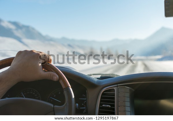 Close-up of\
a man\'s hand on the steering wheel of a car that moves on a snowy\
road among the mountains in the\
winter