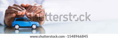 Close-up Of A Man's Hand Protecting The Miniature Blue Car On Reflective White Desk
