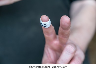 Close-up Of A Man's Hand Showing A Gesture Of Victory. White Armband On Adult Male Index Finger. Smiling Face Painted On The Armband. Positive Emotion Concept. Indoor. Selective Focus.