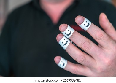 Close-up Of A Man's Hand Showing His Hands To The Camera. White Armbands On Index, Middle, Ring Finger, And Pinky Finger Of Adult Male Hand. Smiling Faces Painted On The Armbands. Helping Hand Concept