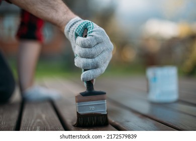 Close-up, a man's hand in a work glove with a painting brush paints boards outdoors. A hand applies paint, oil or varnish to the veranda floorboards