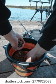 Close-up of a man's hands preparing fish bait and groundmix in an orange and black folding bucket. Professional fishing equipment, including rods and a rod pod, are visible on the wooden dock in the background near the lake. Lifestyle photography of the preparation stage for angling.
