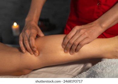 Close-up of a masseur in a red suit massaging the calf muscle of a young woman. Spa massage in a cozy massage therapist's office - Powered by Shutterstock