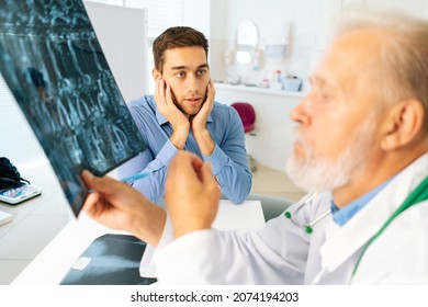 Close-up Of Mature Adult Male Physician Consult Scared Young Man Patient Giving Bad News Explaining Results Of MRI Image. Unhappy Frustrated Young Man Listening To Bad News Sitting In Doctor Office.