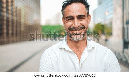 Close-up mature businessman with neat beard is looking at the camera in the financial district in the city. Successful man smiling at the camera on modern buildings background