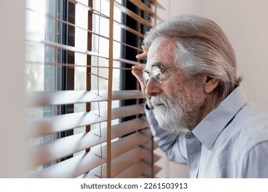 Closeup of mature caucasian man looking through window blinds. Curious elderly man peeking through the blinds by the window. Portrait of thoughtful male observing man watching through window blinds. - Powered by Shutterstock