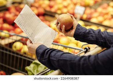 Close-up of mature female customer hands holding shopping list and fresh ripe red apple while choosing fruits in grocery department - Powered by Shutterstock