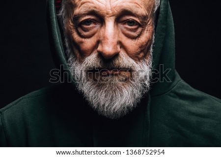 Close-up of a mature old-aged man in green hoodie with wrinkled weathered face, puffy sad eyes looking at camera with mournful expression