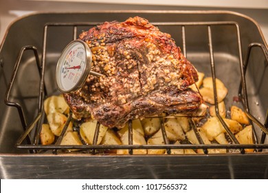 A Closeup Of A Meat Thermometer And A Classic, Succulent Roast Of Beef Covered With Chopped Garlic And Herbs On A Steel Rack In A Large Roasting Pan Filled With Roast Potatoes.