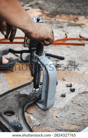 Close-up of a mechanic using a C-clamp press tool to press out a bushing from a car control arm on a workshop floor. Automotive repair, suspension maintenance, and heavy-duty tool concept.