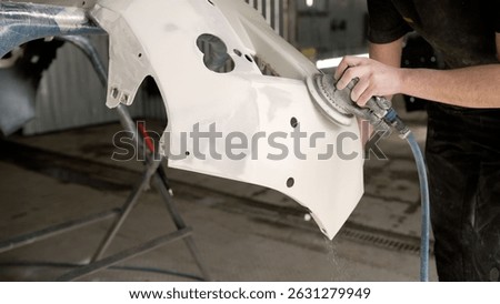 Close-up of mechanic using pneumatic sander, smoothing white car bumper in professional body shop