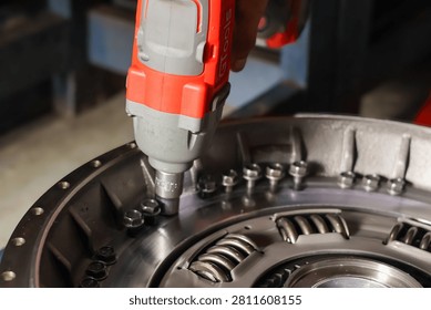 Close-up of a mechanic using a red cordless electric impact wrench to tighten or remove bolts on a heavy vehicle clutch pressure plate assembly in a workshop.