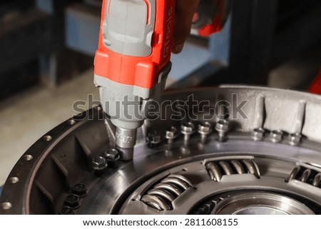 Close-up of a mechanic using a red cordless electric impact wrench to tighten or remove bolts on a heavy vehicle clutch pressure plate assembly in a workshop.