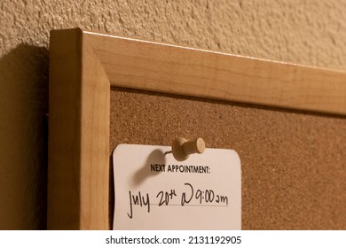 Closeup Of A Medical Appointment Reminder Card With Hand-written Date And Time On A Framed Cork Board At Home.