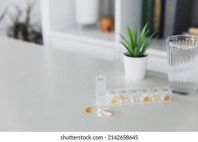 Closeup of medical pill box with doses of tablets for daily take a medicine for treatment, cure the disease. Glass cup of water on table, gray background. Green flowerpot or plant. Helathy lifestyle