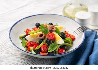 Close-up Of Mediterranean Salad With Anchovy, Green Beans, Tomatoes, Eggs, Black Olives, Capers On White Plate And White Wooden Background. Healthy Food, No Carb Diet Concept.