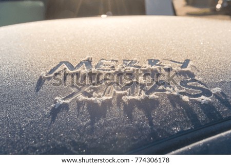 Close-up Merry Christmas (Xmas) handwritten text on top of frosty car. Winter Holidays and Vacations concept. Elegant Merry Xmas and Happy New Year snowy inscription theme. Natural backlit warm light
