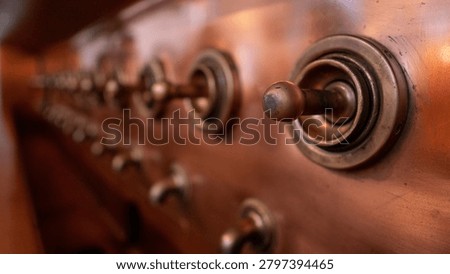 Close-up of metal control knobs mounted on a polished copper brewing vessel inside a brewery. The row of round metal fittings and handles extends along the curved copper surface, which reflects warm ambient lighting. The shallow depth of field emphasizes the foreground control knob while the remaining hardware fades into a soft blur along the vessels surface. The image highlights industrial craftsmanship, mechanical controls, and traditional brewing equipment used in beer production.