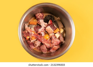 Closeup metal pet bowl with chicken necks, beef meat and bones, paunch, raw egg and carrot pieces. Natural organic daily meal ration preparation for dogs concept. Yellow background, copy space. - Shutterstock ID 2132398303