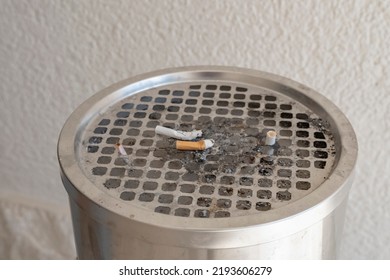 A Close-up Of A Metal Trash Can With An Ashtray Filled With Gobies And Smoked Cigarettes. A City Garbage Can Contaminated With Cigarettes Against A White Wall.