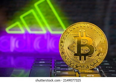 a close-up of a metallic gold coin of the Bitcoin cryptocurrency standing on a laptop keyboard against the background of the Adidas logo