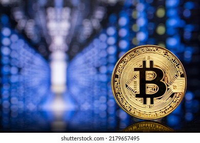 Close-up of a metallic golden Bitcoin cryptocurrency coin standing on a computer board against the backdrop of the colorful lights of a mining farm