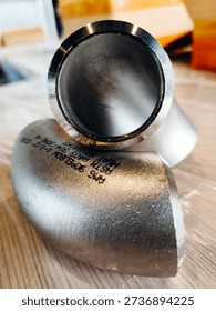 Close-up of a metallic pipe elbow fitting on a wooden surface, showing industrial details and engraved markings