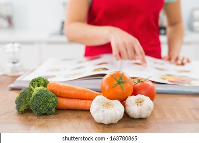 Close-up mid section of a woman with recipe book and vegetables in the kitchen at home