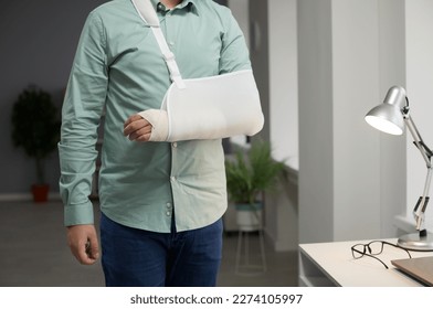 Close-up of the middle part of man with broken bandaged arm in cast. Adult man dressed in casual clothes is standing in the living room with an orthosis or retainer for an injured hand. - Powered by Shutterstock