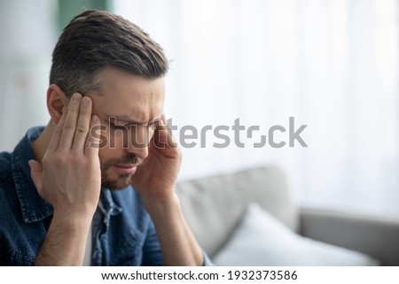 Closeup of middle-aged bearded man suffering from headache at home, touching his temples, panorama with copy space. Migraine, headache, stress, tension problem, hangover concept