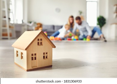 Close-up of miniature detached house on floor at home with happy family playing with little child in background. Mortgage, real estate business, buying property and confidence about future concepts