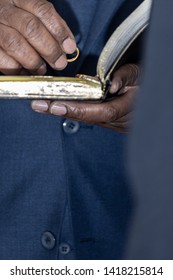 Closeup Of Minister Holding Gold Wedding Ring Above An Open Bible 