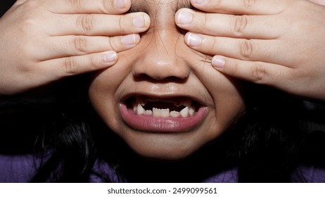close-up of a missing tooth and a small child's face and eyes covered with both hands.