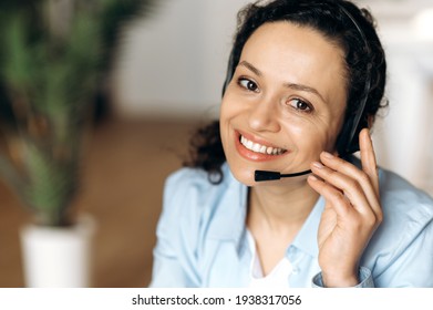 Close-up of mixed race attractive confident adult woman in headset, sitting in office, working as operator of call center or support service, looking directly at the camera and smiling friendly