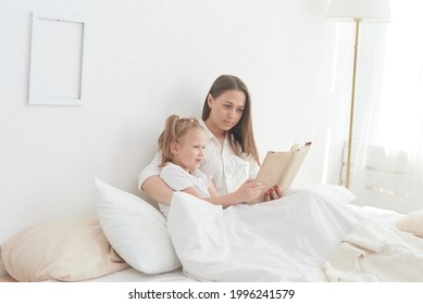 Close-up Mom And Little Daughter Are Resting In A Cozy Bed, Reading A Funny Interesting Children's Book Together