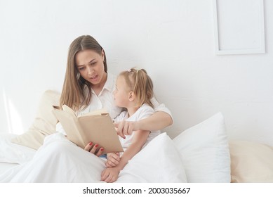 Close-up Mom And Little Daughter Are Resting In A Cozy Bed, Reading A Funny Interesting Children's Book Together