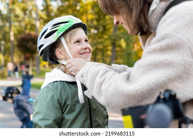 Close-up Mom Parent Hand Help Put On And Fasten Safety Helmet On Cute Blond Caucasian Girl Riding Bike Or Scooter City Street Park Outdoors On Autumn Fall Day. Child Sport Activity Protection Care