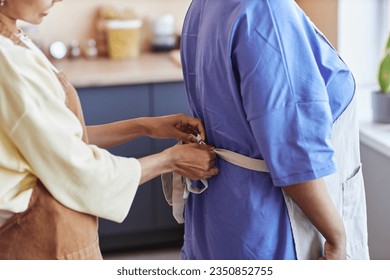 Closeup of mother and daughter tying aprons while cooking together in cozy kitchen, copy space - Powered by Shutterstock