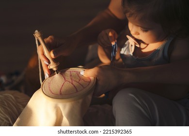 Close-up Of A Mother Showing Her Baby Daughter How To Draw On The Cloth She Is Going To Embroider. Dark Portrait. Family Creative Hand-made Hobby And Leisure