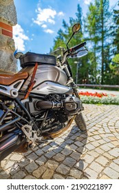 Closeup Motorcycle Parked On Driveway Road In Warm Sunny Light. Blurred Forest Trees, Park Flowers And Gravel Stone Road. Classic Vintage Motorbike, Outdoor Sport Recreational Pursuit. Nature Travel