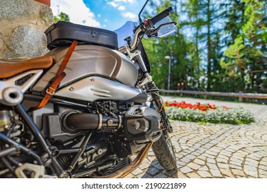 Closeup Motorcycle Parked On Driveway Road In Warm Sunny Light. Blurred Forest Trees, Park Flowers And Gravel Stone Road. Classic Vintage Motorbike, Outdoor Sport Recreational Pursuit. Nature Travel