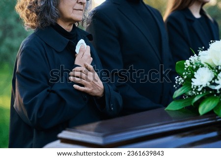 Close-up of mourning mature woman in black attire keeping hands on chest while standing next to her family during funeral at graveyard