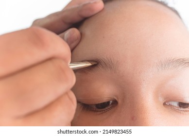 Closeup Of A MUA Or Makeup Artist Applying Brow Filler With A Slanted Eyeliner Brush On The Eyebrow Of A Young Female Asian Model.