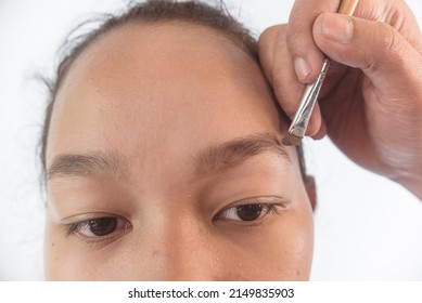 Closeup of a MUA or Makeup artist using brow filler on a slanted eyeliner brush to shape the eyebrow of a young female asian model. Early stages of makeup.