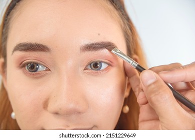 Closeup of a MUA or Makeup artist using brow filler on a slanted eyeliner brush to shape the eyebrow of a young female asian model.