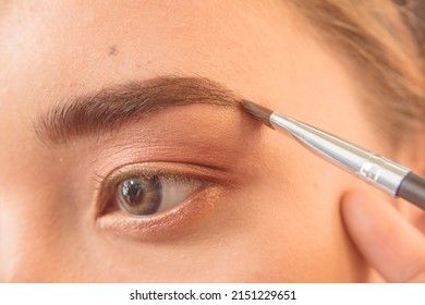 Closeup of a MUA or Makeup artist using brow filler on a slanted eyeliner brush to shape the eyebrow of a young female asian model. Early stages of makeup.