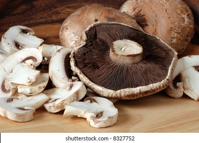 Closeup Of Mushrooms.  Large Portabello Mushrooms Arranged On A Wooden Cutting Board With Smaller Sliced Mushrooms.  Wood Grain Bowl In Soft Focus As Background.