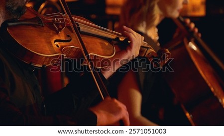 A close-up of musicians playing string instruments in an orchestra, focusing on a violinist and a cellist. The warm lighting highlights the wood of the instruments