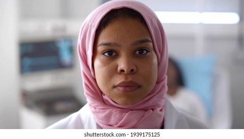 Close-up of muslim female doctor looking at camera working at hospital with patient lying in bed on background. Arabian female medical worker in hijab posing at camera