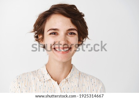 Close-up of natural cute girl smiling with white teeth, looking happy and excited at camera, standing on white background.